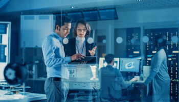 In Technology Research Facility: Female Project Manager Talks With Chief Engineer, they Consult Tablet Computer. Team of Industrial Engineers, Developers Work on Engine Design Using Computers